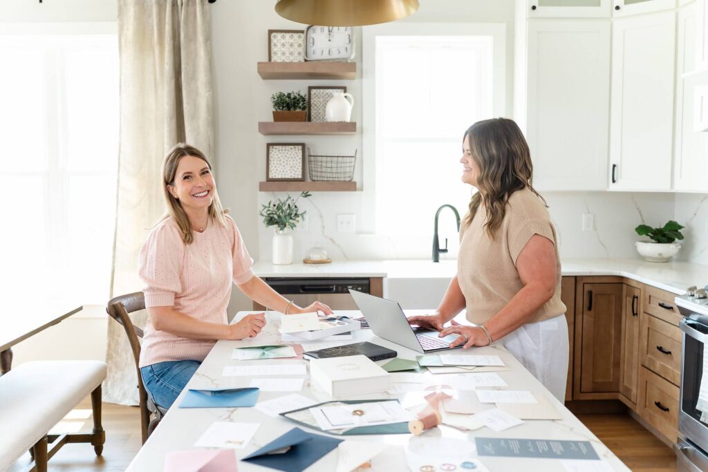 Wedding stationers work at a computer surrounded by their stationery designs on a kitchen island