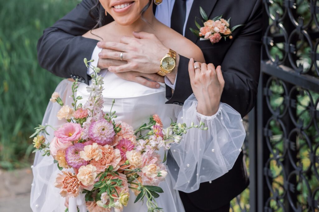 Closeup of a couple on their romantic and timeless summer garden party wedding at Tyler Gardens. Closeup image of the groom's arms around the bride and bride is holding bridal bouquet featuring soft peach, pink, and greenery. 