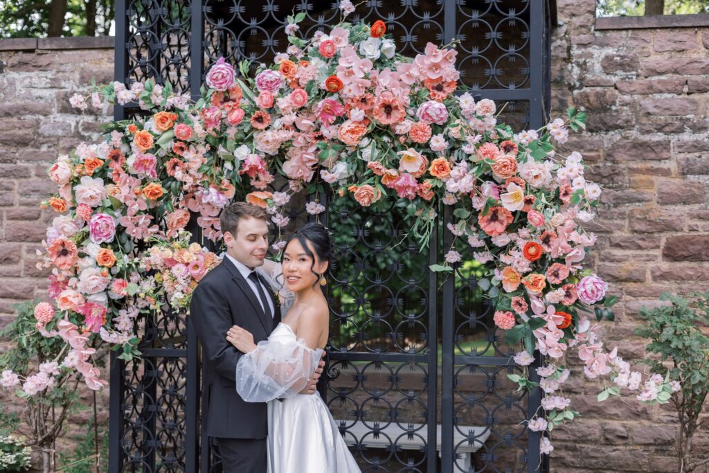 Newly married couple posing together on their wedding day at Tyler Gardens. Full flower arch surrounds the couple and features accents of peach, pink, yellow, orange, and greenery.