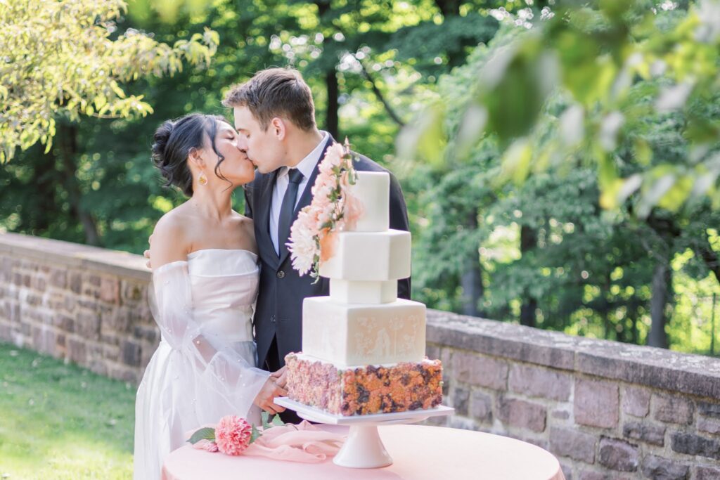 Couple kissing on their wedding day at Tyler Gardens and they are standing next to their wedding cake which picks up on peach accents to build off of the summer garden party theme.