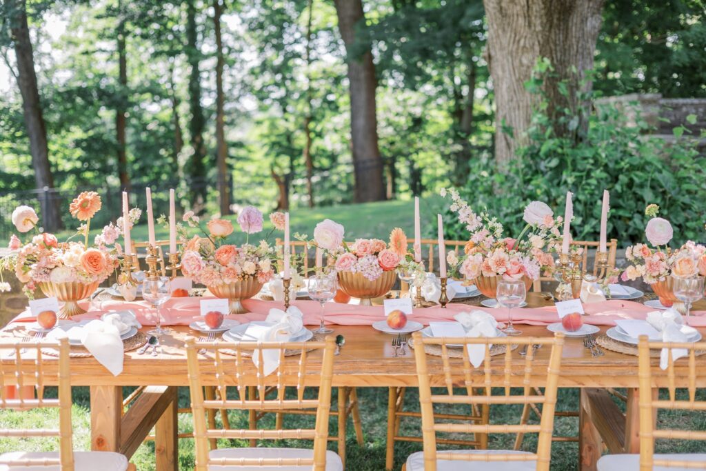 Full table setup at Tyler Gardens summer garden party wedding. Features Pantone's color of the year, "Peach Fuzz." Candlesticks with gold accents and full floral centerpieces in peach tones. Escort cards are placed in fresh peaches next to each place setting.