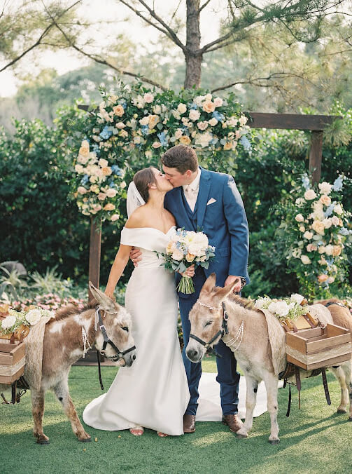 Beer donkeys stand next to bride and groom who are kissing under a ceremony arch