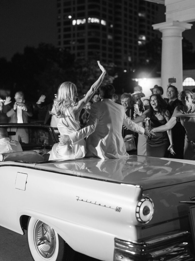 Bride and groom exiting their reception in a classic car as guests cheer them on