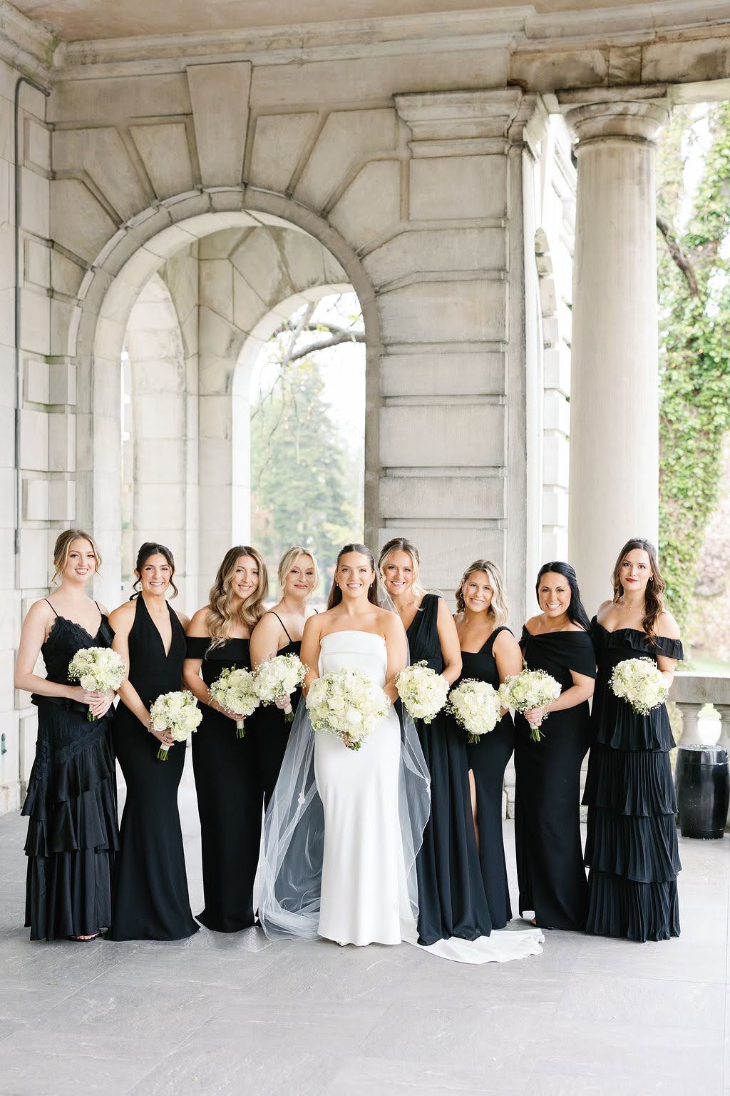 Bride with eight bridesmaids all dressed in unique formal black dresses with white bouquets
