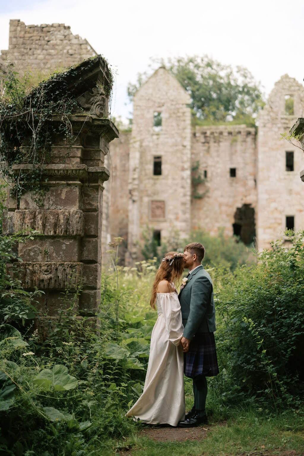 Groom wearing traditional kilt kisses bride in front of Dalquharran Castle