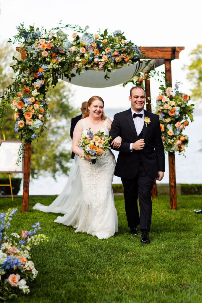 Bride and groom walk down the aisle arm in arm in front of a chuppah adorned with colorful flowers