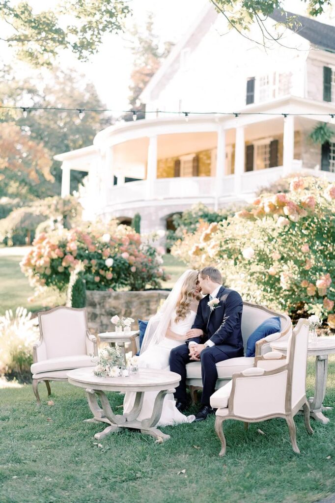 Bride and groom cuddle on an elegant outdoor lounge sofa, surrounded by a table and chairs