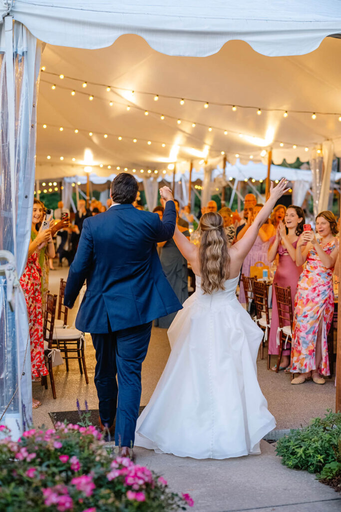 Bride and groom walk into a wedding reception tent as guest cheer them on