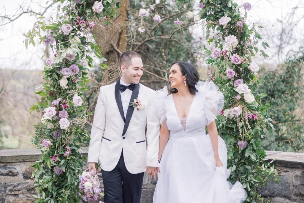 Bride and groom smile at each other in front of an outdoor ceremony arch with purple flowers