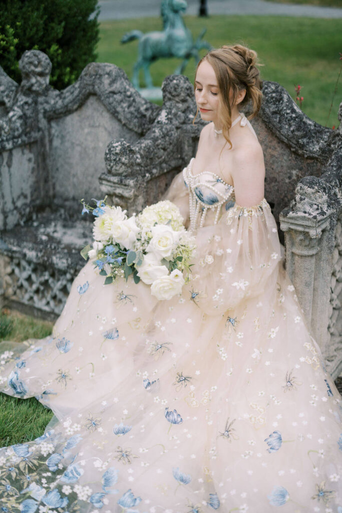 Bride sits upon a stone bench wearing an elegant bridal gown adorned with pearls and blue flowers