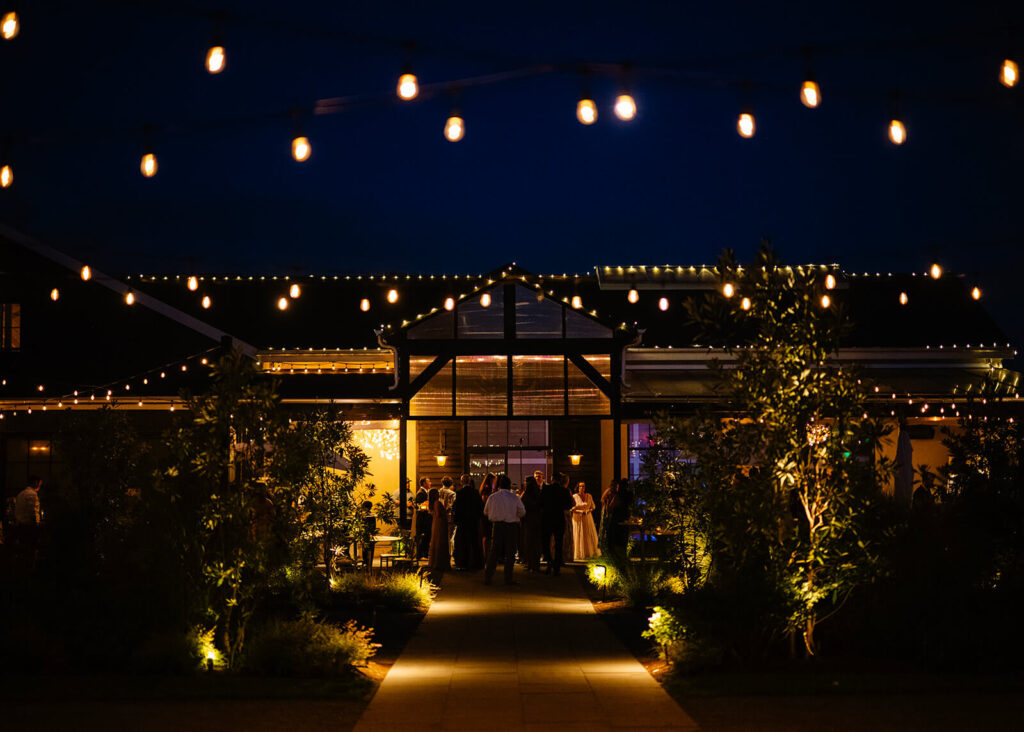 Exterior of Terrain Gardens at DelVal at night as guests mingle on the patio during a wedding reception