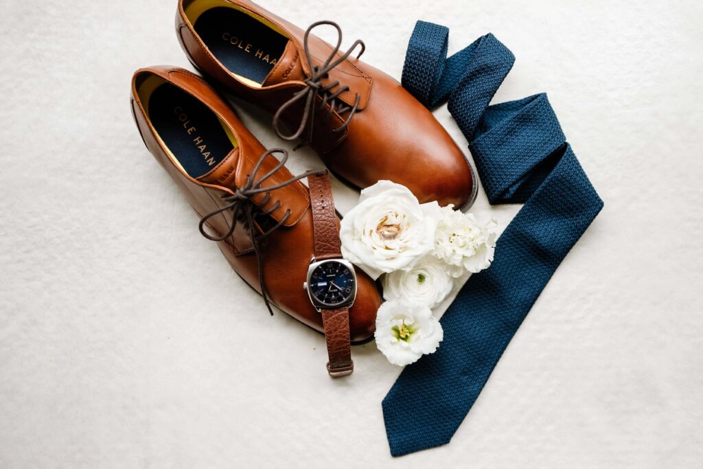Wedding day accessories belonging to the groom, like shoes, a watch, and a blue tie