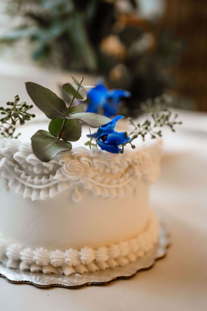 Elegant and ornate white ceremonial cutting cake with greenery and blue flowers on top