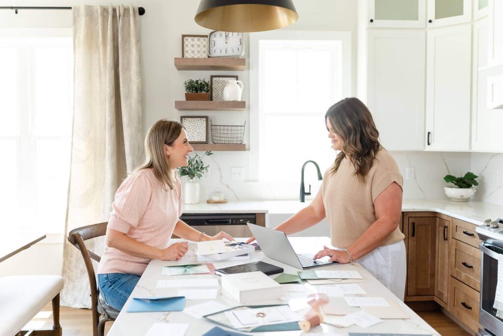 Heather and Emily, co-owners of Heather Paperie, designing custom wedding invitations at a kitchen island filled with stationery inspiration