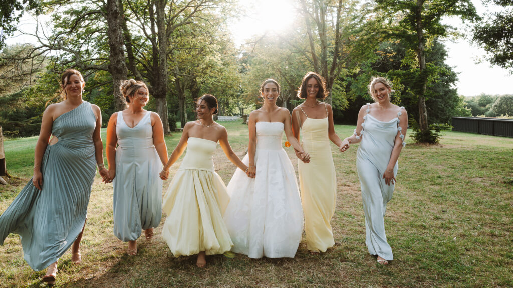 Bride walking through a shaded grove with bridesmaids, wearing light blue and yellow