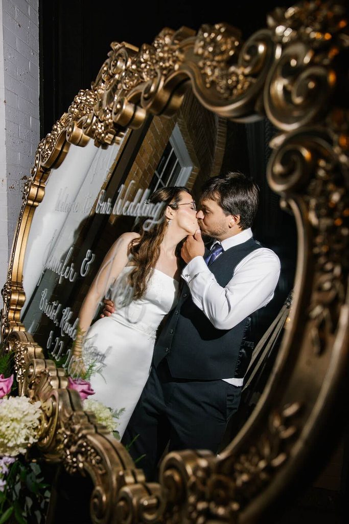 Bride and groom seen kissing reflected in the glass of an ornate gold mirror