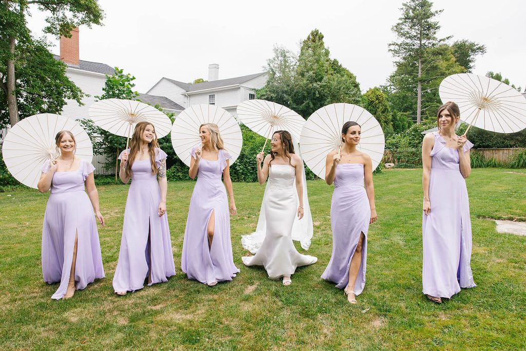 Bridesmaids, dressed in light purple, walking with the bride and  holding parasols